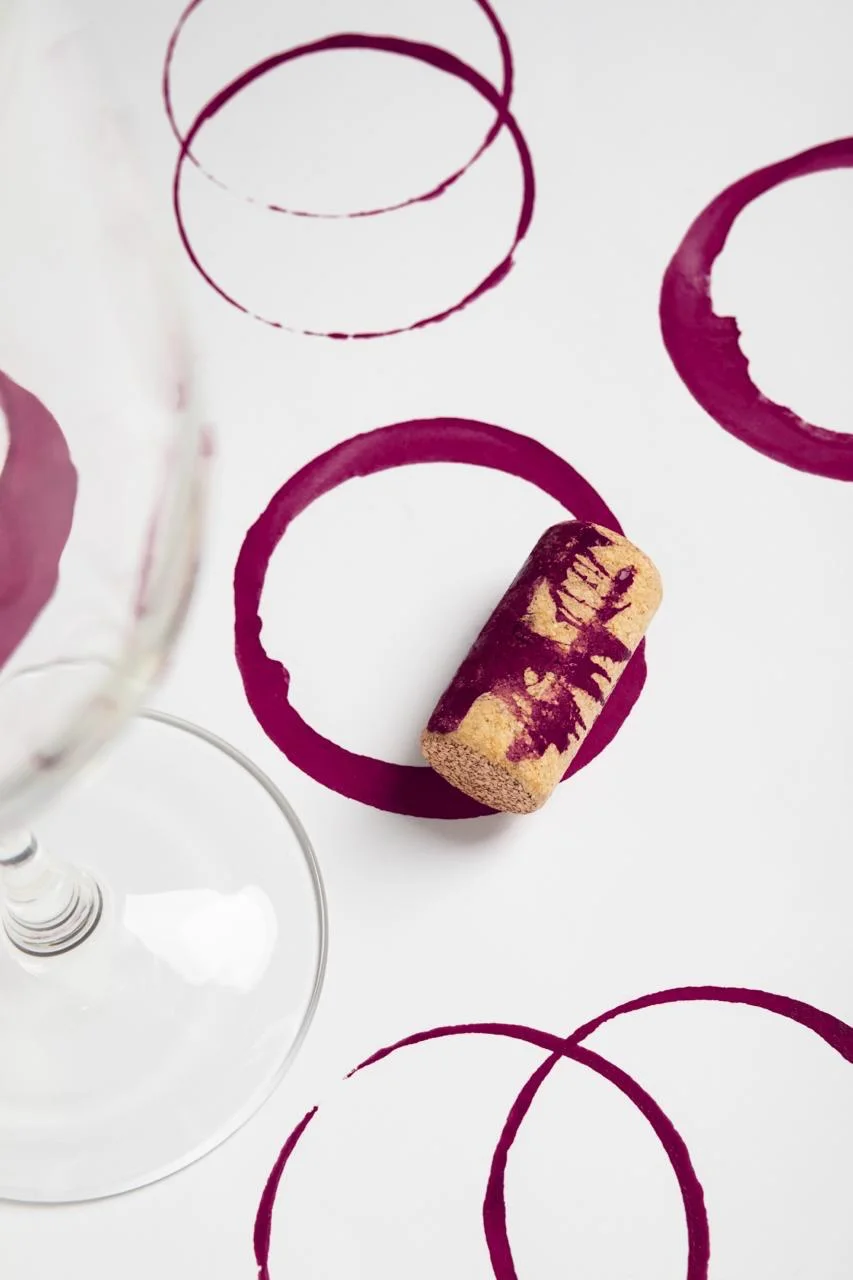 A glass of red wine beside a cork, with wine rings on a white surface
