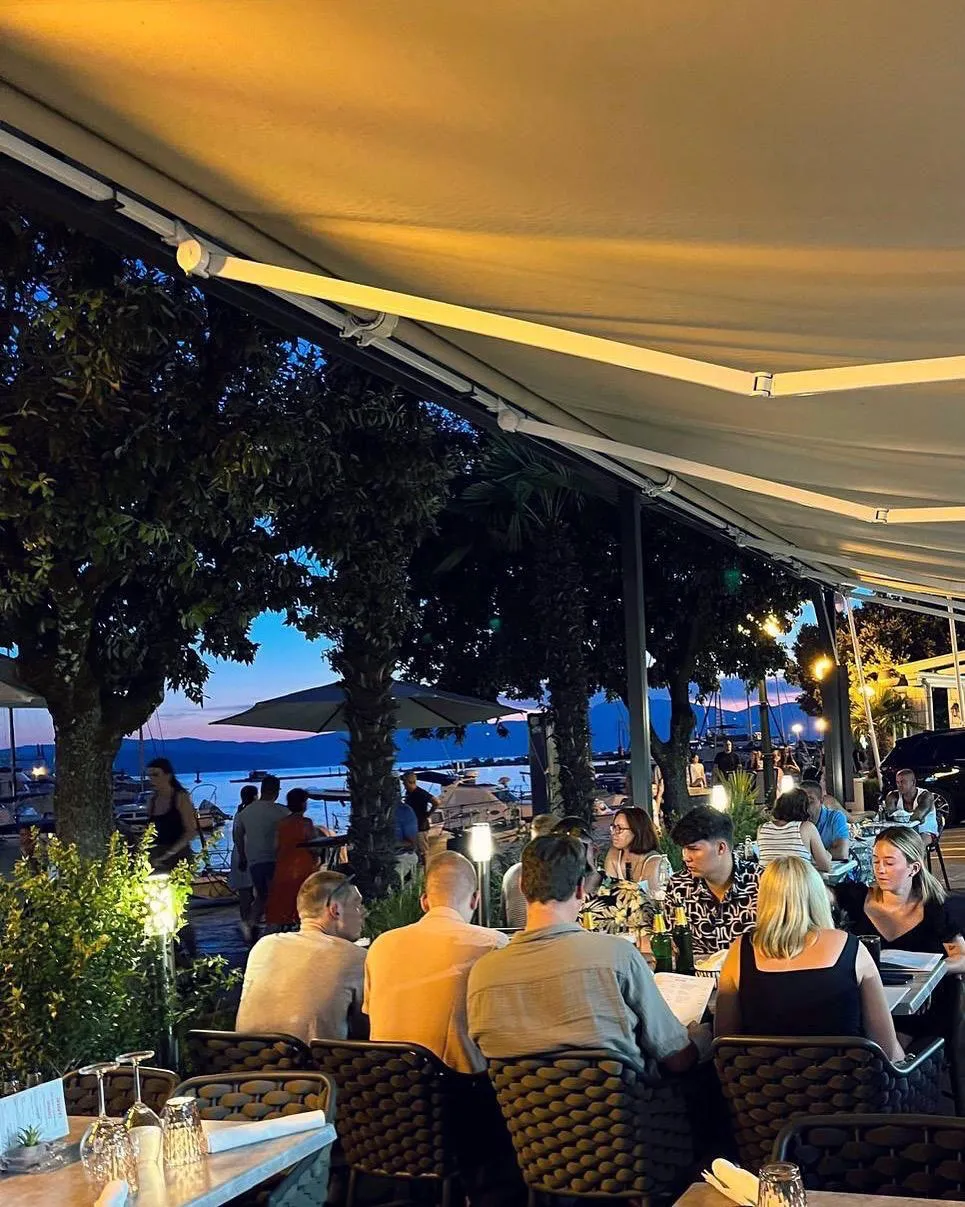 Guests dining on the seafront terrace at dusk, with the Malinska harbour beyond