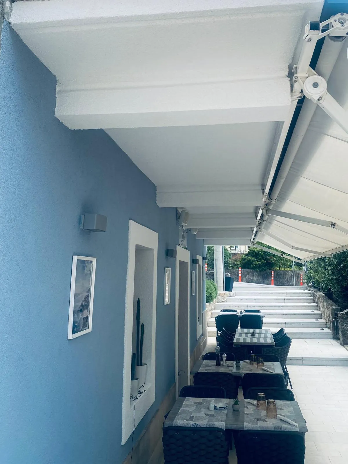 The restaurant's covered terrace — a blue-washed wall with framed art and set tables under a white awning, beside the stone steps